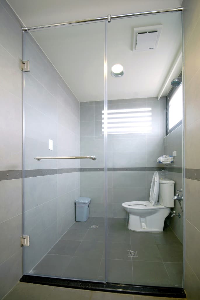 Baño moderno con puerta de vidrio transparente, azulejos grises y sanitarios blancos.