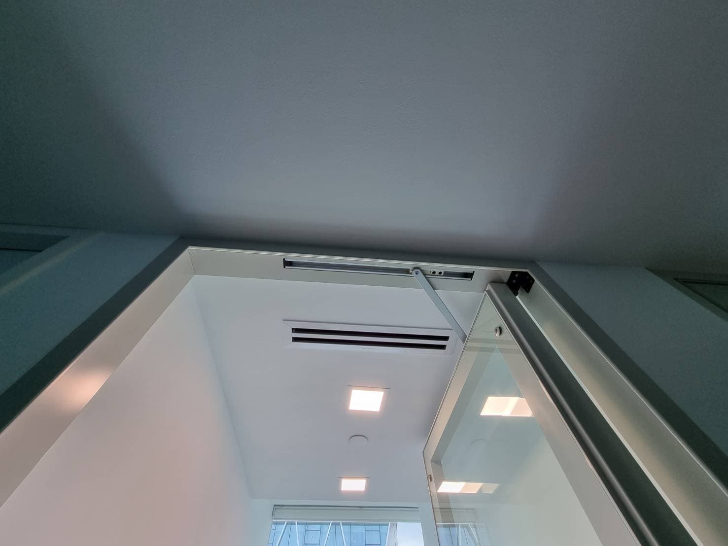 仰视半开的银框玻璃门,上方装有闭门器,背景是带灯和通风口的白色天花板。